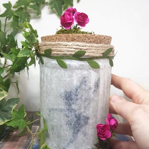 Puede incluir: Un tarro blanco con tapa de corcho decorado con flores rosas, hojas verdes y cuerda. El tarro tiene un estilo r&uacute;stico y de granja.
