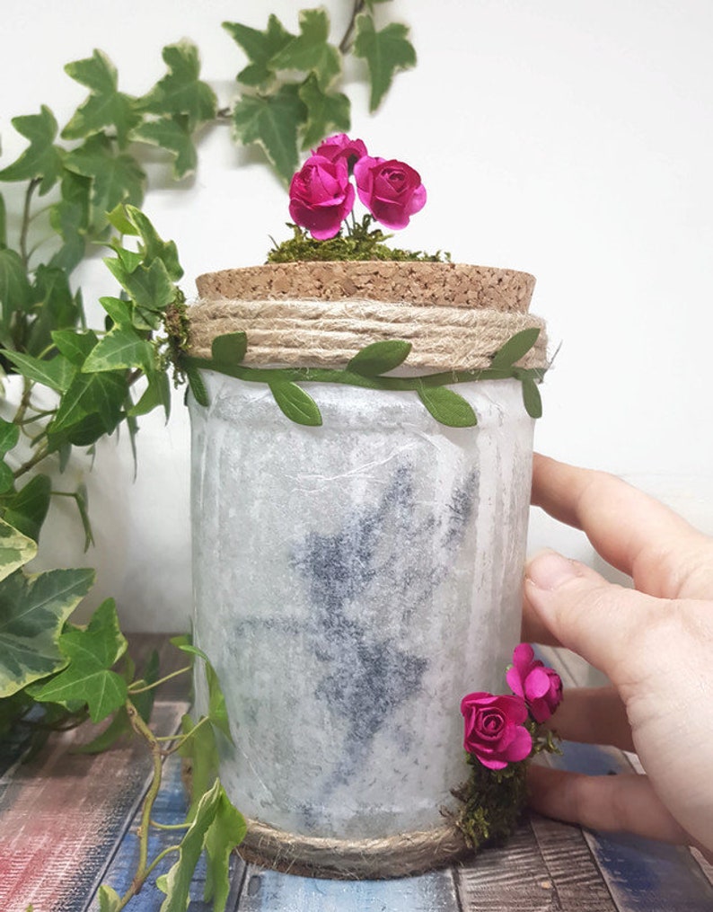 Puede incluir: Un tarro blanco con tapa de corcho decorado con flores rosas, hojas verdes y cuerda. El tarro tiene un estilo r&uacute;stico y de granja.