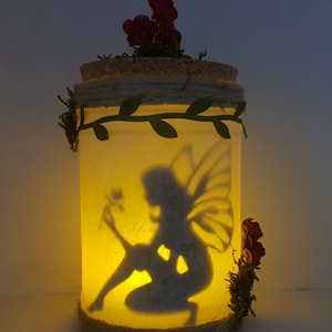 May include: A glass jar with a cork lid and a silhouette of a fairy holding a flower inside. The jar is decorated with a green vine and red flowers.