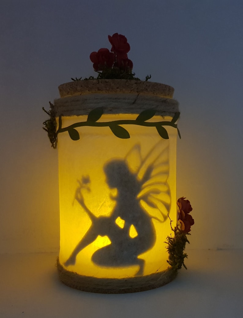 May include: A glass jar with a cork lid and a silhouette of a fairy holding a flower inside. The jar is decorated with a green vine and red flowers.