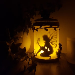 May include: A glass jar with a fairy silhouette inside, illuminated by a candle. The jar is decorated with leaves and has a rustic twine lid. The fairy is holding a flower and has wings.