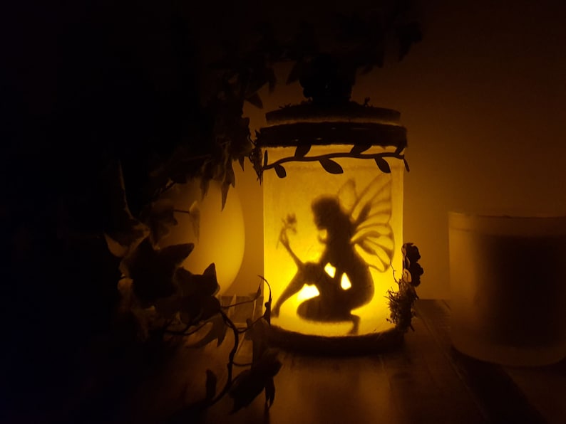 May include: A glass jar with a fairy silhouette inside, illuminated by a candle. The jar is decorated with leaves and has a rustic twine lid. The fairy is holding a flower and has wings.