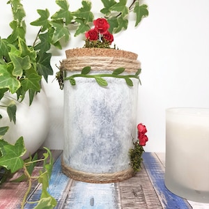 May include: A white ceramic jar with a cork lid, decorated with twine, faux greenery, and red flowers. The jar is sitting on a blue and white wooden surface.
