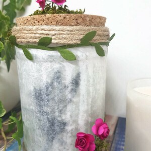 Puede incluir: Un tarro de vidrio blanco con tapa de corcho, decorado con cuerda, hojas verdes y rosas de papel rosa. El tarro est&aacute; sobre una superficie de madera azul y blanca.