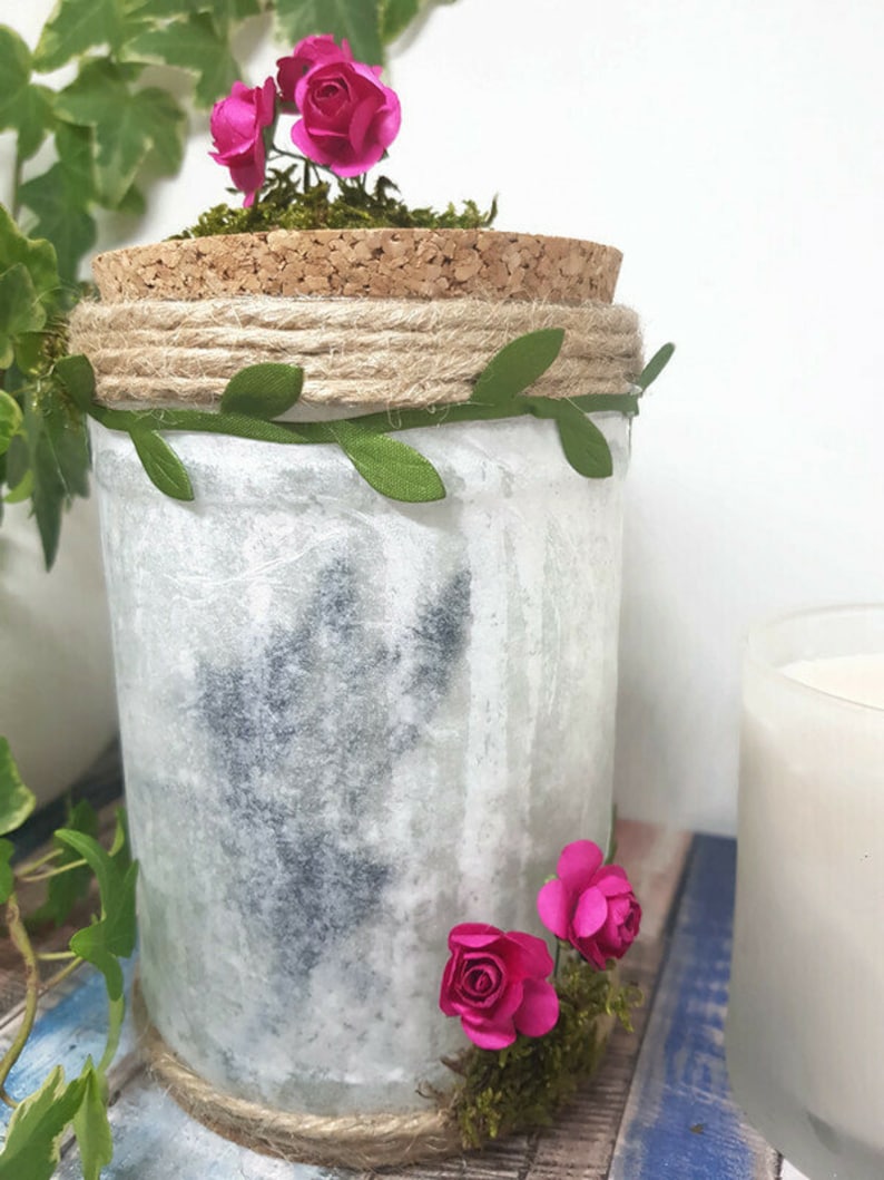 Puede incluir: Un tarro de vidrio blanco con tapa de corcho, decorado con cuerda, hojas verdes y rosas de papel rosa. El tarro est&aacute; sobre una superficie de madera azul y blanca.