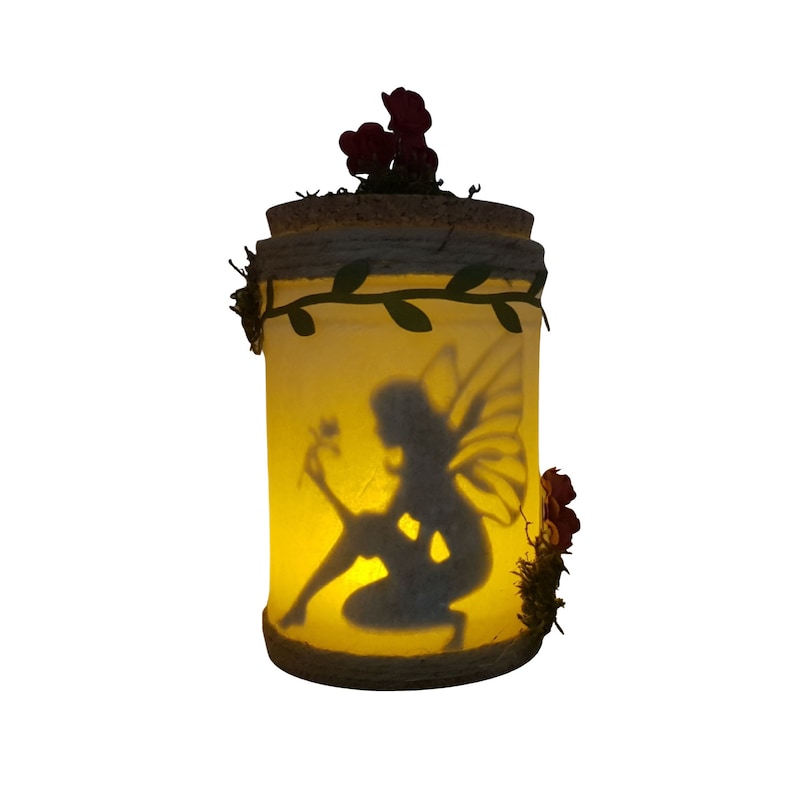 May include: A glass jar with a fairy silhouette inside. The jar is decorated with a brown cork lid, a green vine, and pink flowers.