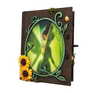 May include: A wooden book with a green and brown leafy design. The book has a round window with a picture of a fairy hidden behind a green leaf. The book is decorated with sunflowers.