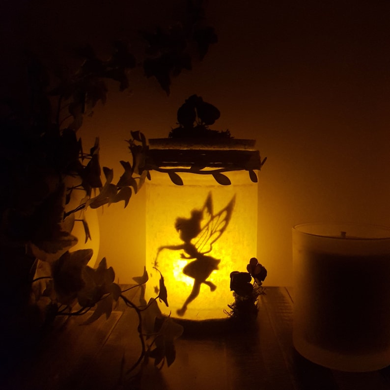 Puede incluir: Un tarro de vidrio con una silueta de hada en su interior, iluminado por una vela. El tarro est&aacute; decorado con hojas y tiene una tapa con musgo en la parte superior. El tarro est&aacute; sentado sobre una superficie de madera junto a una vela blanca.