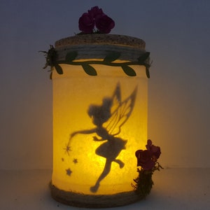Puede incluir: Un tarro de vidrio con una silueta de hada y estrellas en su interior. El tarro est&aacute; decorado con cuerda, hojas y flores moradas.