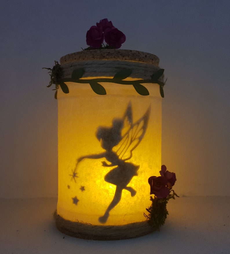 Puede incluir: Un tarro de vidrio con una silueta de hada y estrellas en su interior. El tarro est&aacute; decorado con cuerda, hojas y flores moradas.