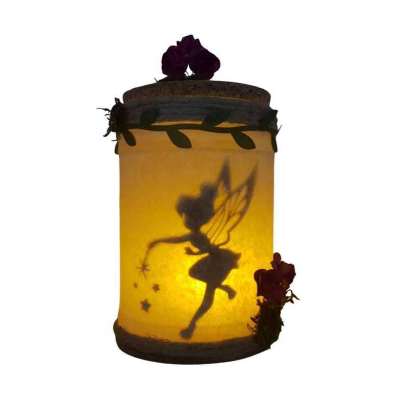Puede incluir: Un tarro de vidrio con una silueta de hada y un brillo en el interior. El tarro est&aacute; decorado con una tapa de corcho marr&oacute;n, hojas verdes y flores moradas.