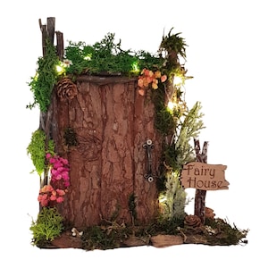 Może przedstawiać: Miniaturowy domek dla wróżek z drewnianymi drzwiami, ozdobiony mchem, kwiatami i lampkami. Mały znak głosi "Fairy House". Drzwi mają rustykalną fakturę kory i metalową klamkę. Fantazyjny element dekoracyjny.