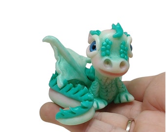 May include: A small, hand-crafted, green and white dragon figurine with blue eyes. The dragon is sitting on a hand with its wings spread out.