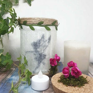Puede incluir: Un tarro de vidrio blanco con un dise&ntilde;o de silueta de hada, decorado con cuerda y hojas verdes. El tarro est&aacute; iluminado con una vela de t&eacute; y rodeado de flores de papel rosa y musgo sobre una base de corcho.