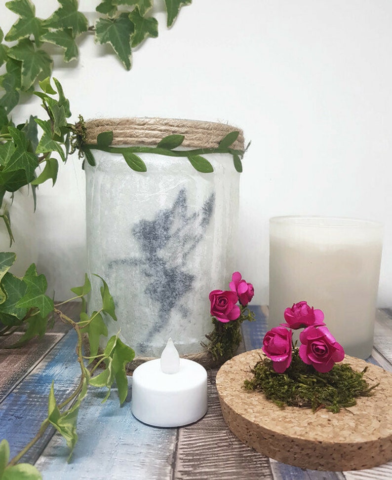 Puede incluir: Un tarro de vidrio blanco con un dise&ntilde;o de silueta de hada, decorado con cuerda y hojas verdes. El tarro est&aacute; iluminado con una vela de t&eacute; y rodeado de flores de papel rosa y musgo sobre una base de corcho.