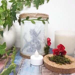 May include: A white glass jar with a silhouette of a fairy inside, decorated with a green vine and twine around the top. The jar is sitting on a wooden surface with a small white tea light and a cork lid with moss and red roses.