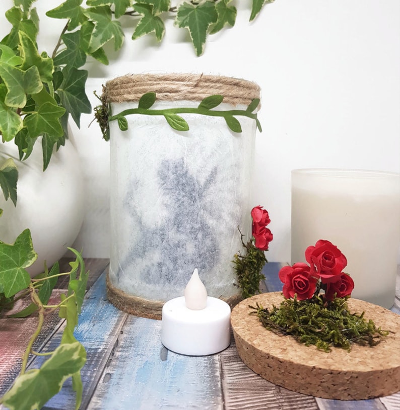May include: A white glass jar with a silhouette of a fairy inside, decorated with a green vine and twine around the top. The jar is sitting on a wooden surface with a small white tea light and a cork lid with moss and red roses.