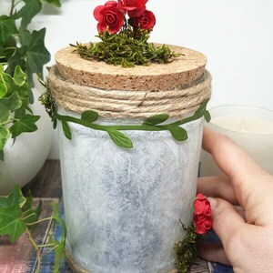 May include: A white glass jar with a cork lid decorated with moss, red paper roses, and twine. The jar is wrapped with a green leaf garland and has a rustic, farmhouse aesthetic.