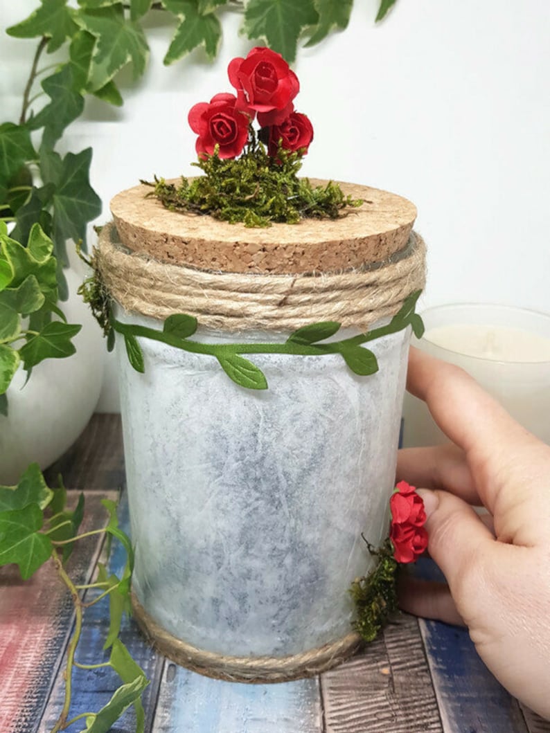 May include: A white glass jar with a cork lid decorated with moss, red paper roses, and twine. The jar is wrapped with a green leaf garland and has a rustic, farmhouse aesthetic.