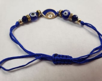 May include: A blue and gold evil eye bracelet with a charm in the center. The bracelet is made of string and has a knot at the end.