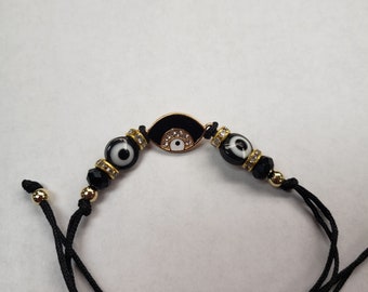May include: Black string bracelet with a gold evil eye charm and black beads. The charm has a white eye with a black pupil and is surrounded by clear rhinestones.