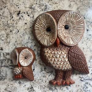 May include: Two brown ceramic owls with white eyes and brown and white patterned feathers. The larger owl is about 25 cm tall and the smaller owl is about 12 cm tall.