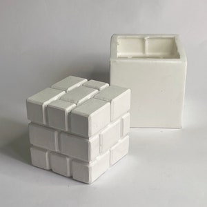 May include: Two white ceramic objects. One is a cube with a brick-like pattern, and the other is a square container. The cube is a solid, three-dimensional form, while the container has an open top and a hollow interior.
