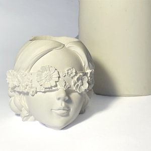 May include: A white, sculpted head-shaped planter with a floral blindfold. The planter has a smooth finish and a hollowed-out top for planting. A cylindrical object is in the background.
