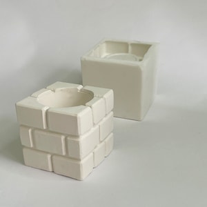 May include: Two white ceramic planters, one shaped like a brick wall and the other a simple cube. Both have a small opening at the top for planting.