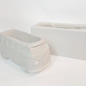 Pode incluir: Um vaso de cerâmica cinza claro em forma de carrinha vintage, com uma abertura retangular. Ao lado, um molde retangular. A carrinha tem rodas, janelas e uma grelha frontal. O vaso é para interior ou exterior.