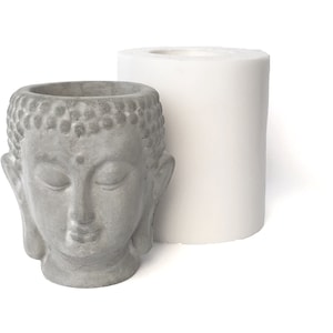 Può includere: Un vaso da fiori in cemento grigio a forma di testa di Buddha con gli occhi chiusi. Il vaso è accanto a uno stampo in silicone bianco.