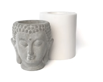 Könnte beinhalten: Ein grauer Betontopf in Form eines Buddha-Kopfes mit geschlossenen Augen. Der Topf steht neben einer weißen Silikonform.