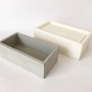Pode incluir: Duas jardineiras retangulares de concreto. Uma é cinza claro, a outra é branco sujo. Ambas têm um design simples e moderno com laterais retas e formato retangular. As jardineiras estão vazias e sobre uma superfície branca.