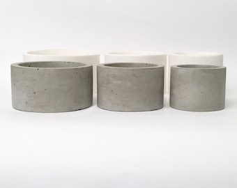 May include: Three concrete planters in different sizes, with a light gray color and a rough texture. The planters are arranged in a row from largest to smallest.