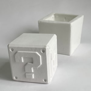 May include: Two white ceramic planters shaped like a question mark block from the Super Mario Bros. video game. The smaller planter has a question mark on the front and the larger planter is open at the top.