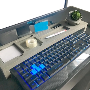 May include: A gray concrete desk organizer with multiple compartments for pens, pencils, tape, and other office supplies. A keyboard with blue backlit keys is placed on the organizer.