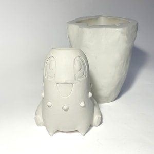 May include: Two unpainted ceramic vases. One is a tall, irregular shape. The other is a small, cartoon-like figure with a smiling face and spiky details. Both are white and sit on a white surface.