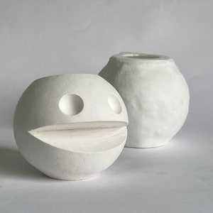 May include: Two white ceramic planters, one shaped like a face with two round eyes and a mouth, the other a round, smooth shape.