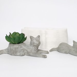 Può includere: Due sculture di vasi per piante a forma di gatto in cemento, una con una pianta grassa verde, su uno sfondo bianco.