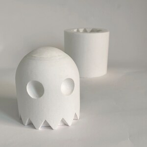 Pode incluir: Vaso de cerâmica branco em forma de fantasma com a parte superior arredondada e a inferior dentada. O fantasma tem dois olhos redondos e uma pequena boca.