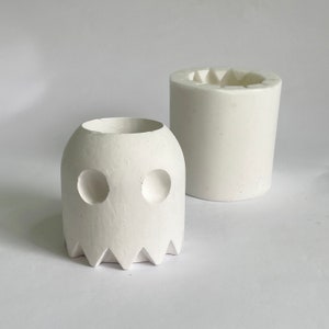 Op de afbeelding: Twee witte keramische potten, een in de vorm van een spook met twee ronde ogen en een gekartelde onderkant, de andere een eenvoudige cilinder met een stervormige bovenkant.