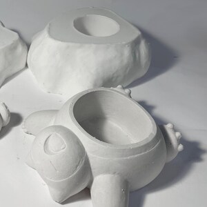 May include: White ceramic planter shaped like a cartoon bear with a round opening for planting. The planter is sitting on a white surface.