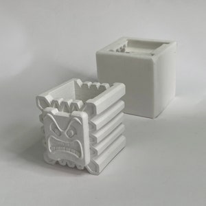 Op de afbeelding: Twee witte keramische potten, een met een vierkante vorm en de andere met een vierkante vorm en een gegraveerd boos gezicht aan de voorkant.