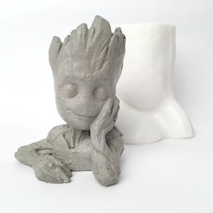 May include: A grey concrete planter shaped like a character from the movie Guardians of the Galaxy. The planter is sitting on a white surface. The planter is a great way to add a touch of whimsy to your home decor.