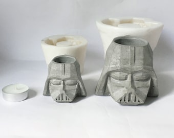 Könnte beinhalten: Zwei graue Beton-Pflanzer in Form von Darth Vaders Kopf. Die Pflanzer sind unterschiedlich groß und haben eine glatte, strukturierte Oberfläche. Eine kleine weiße Teelichtkerze befindet sich im Vordergrund.