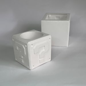 May include: Two white ceramic planters shaped like question mark blocks from the Super Mario Bros. video game. The smaller planter has a question mark embossed on each side.