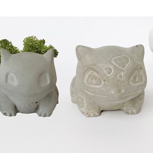 Puede incluir: Tres macetas de hormigón con forma de personajes de Pokémon. La maceta de la izquierda es un Bulbasaur gris claro con musgo verde creciendo en su interior. La maceta del centro es un Squirtle gris claro con un fondo gris claro. La maceta de la derecha es un Charmander blanco con un fondo blanco.