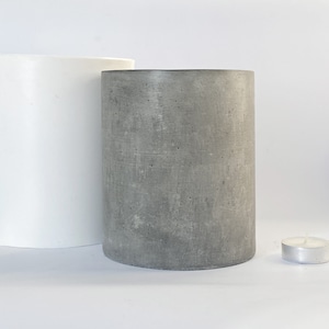 May include: Two cylindrical concrete candle holders, one white and one gray, are displayed. A small, silver tea light candle sits to the right. The candle holders are smooth and modern, suitable for home decor.