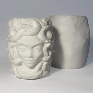 Può includere: Vaso in ceramica bianca a forma di testa di Medusa, con serpenti come capelli. Il vaso è accanto a un vaso cilindrico bianco semplice.