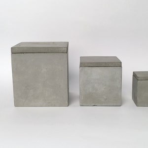 Puede incluir: Tres cajas de hormigón gris de diferentes tamaños, cada una con una tapa plana. Las cajas son cuadradas y tienen un diseño minimalista. La caja más grande mide aproximadamente 25 cm de alto, la mediana 20 cm y la más pequeña 15 cm.